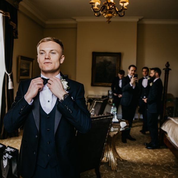 A groom in formal wear adjusts his bow tie, while his bestmen wait patently in the background.