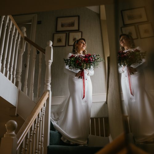 A bride in a white dress stands on a staircase holding a bouquet, with her reflection visible in a nearby mirror.