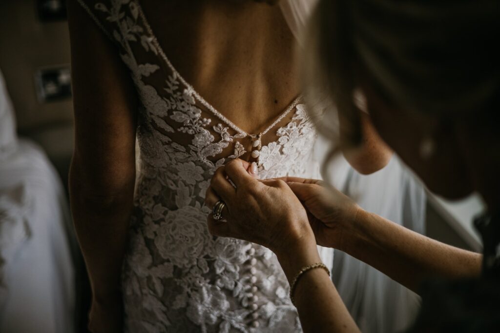 A close-up of a bride getting dressed, with someone fastening the intricate lace back of her wedding gown.