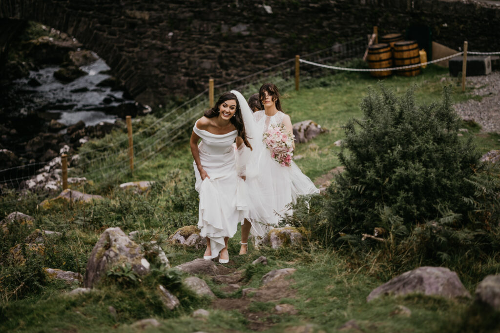 A bride and her bridesmaid walk together on a rocky path by a river, surrounded by lush greenery.
