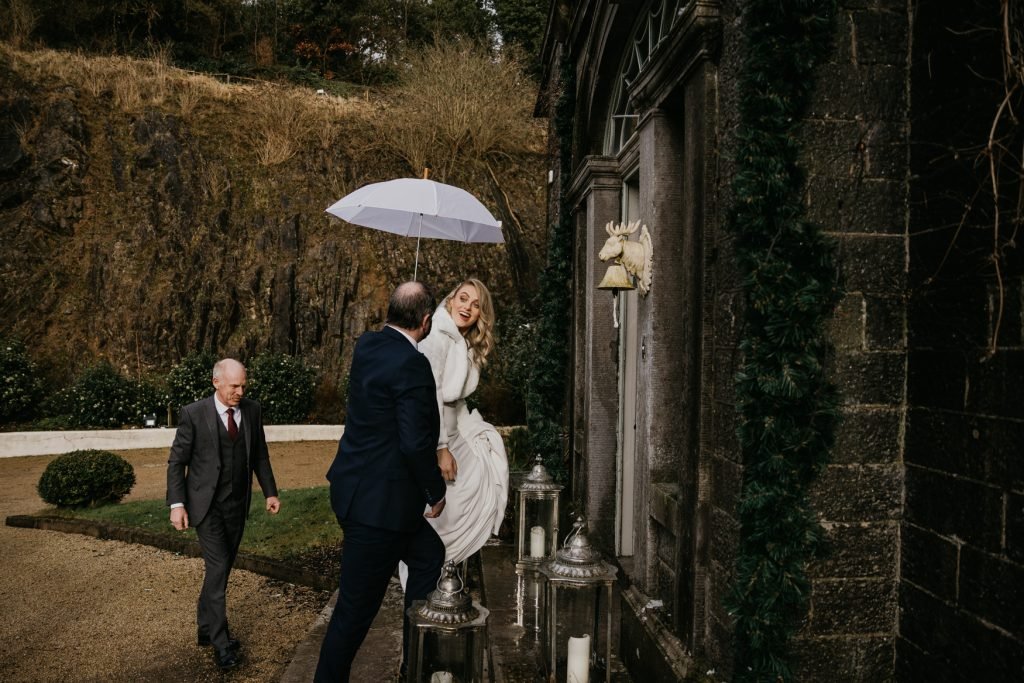 A bride is being assisted by a man while holding an umbrella, with another man walking in the background.