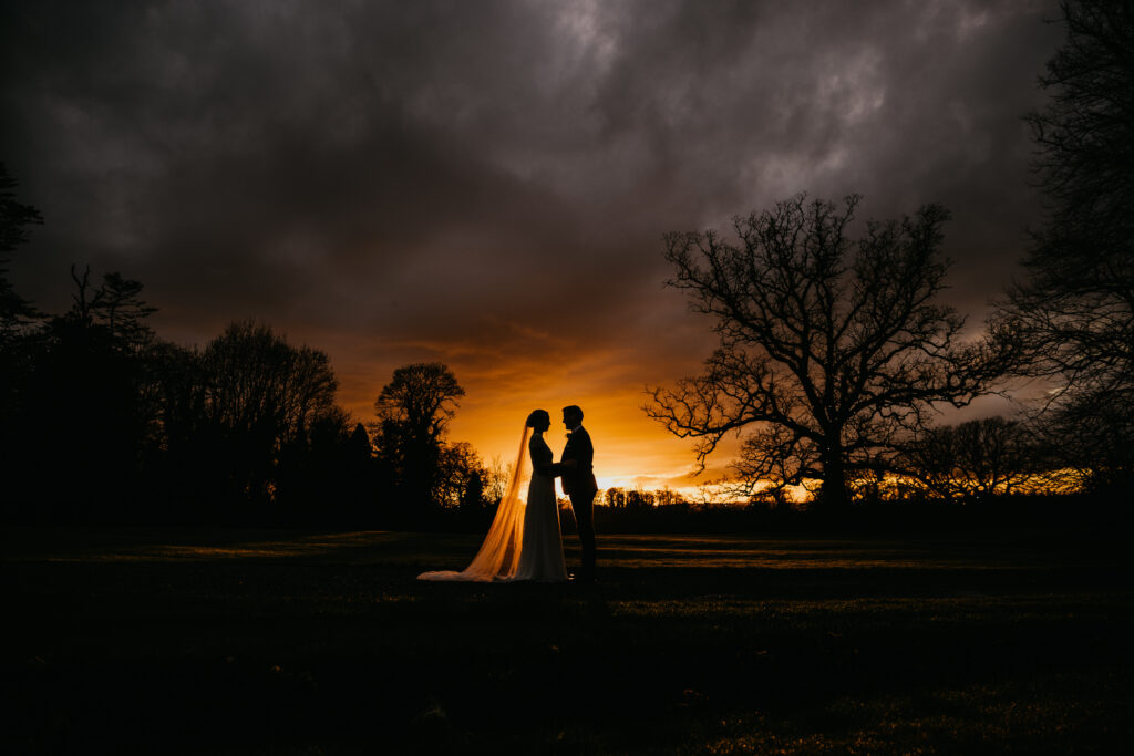 Bride and groom holding hands under a moody, orange sky at sunset.