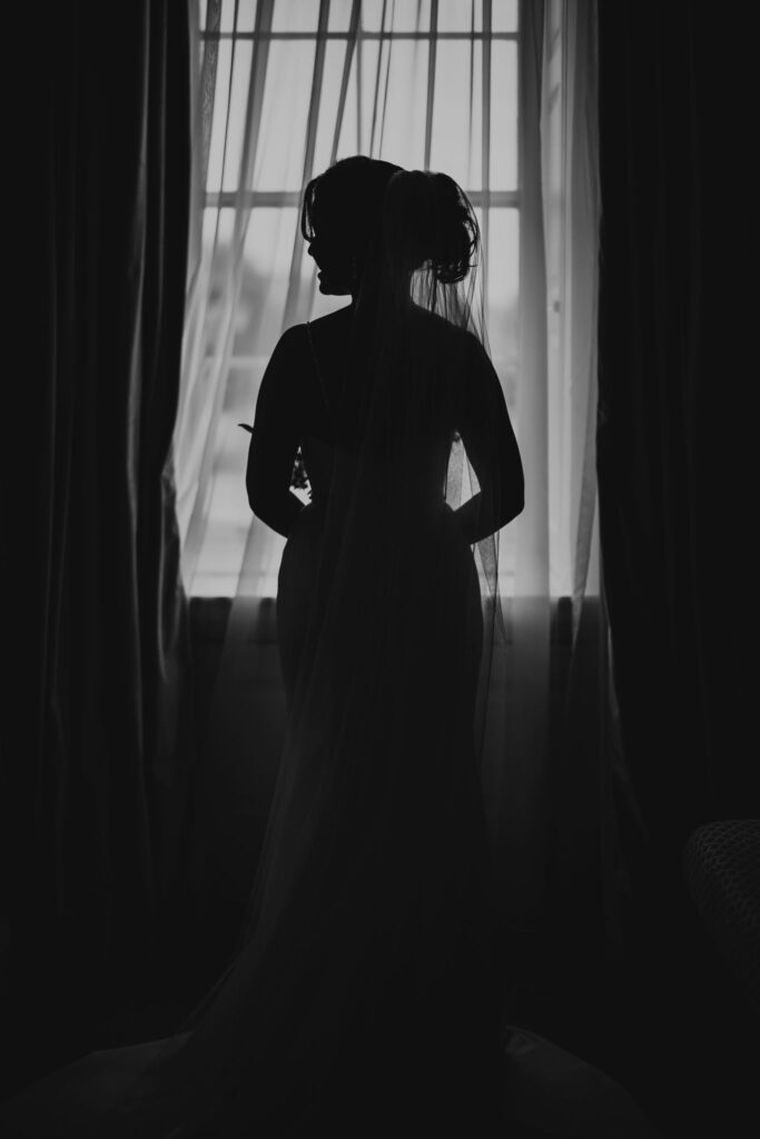 Silhouette of the bride standing before a window with sheer curtains