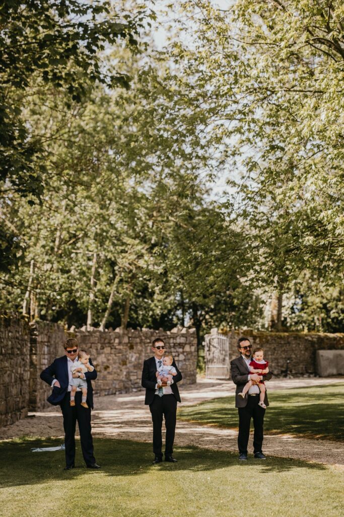Three groomsmen each holding a baby, standing on a lawn