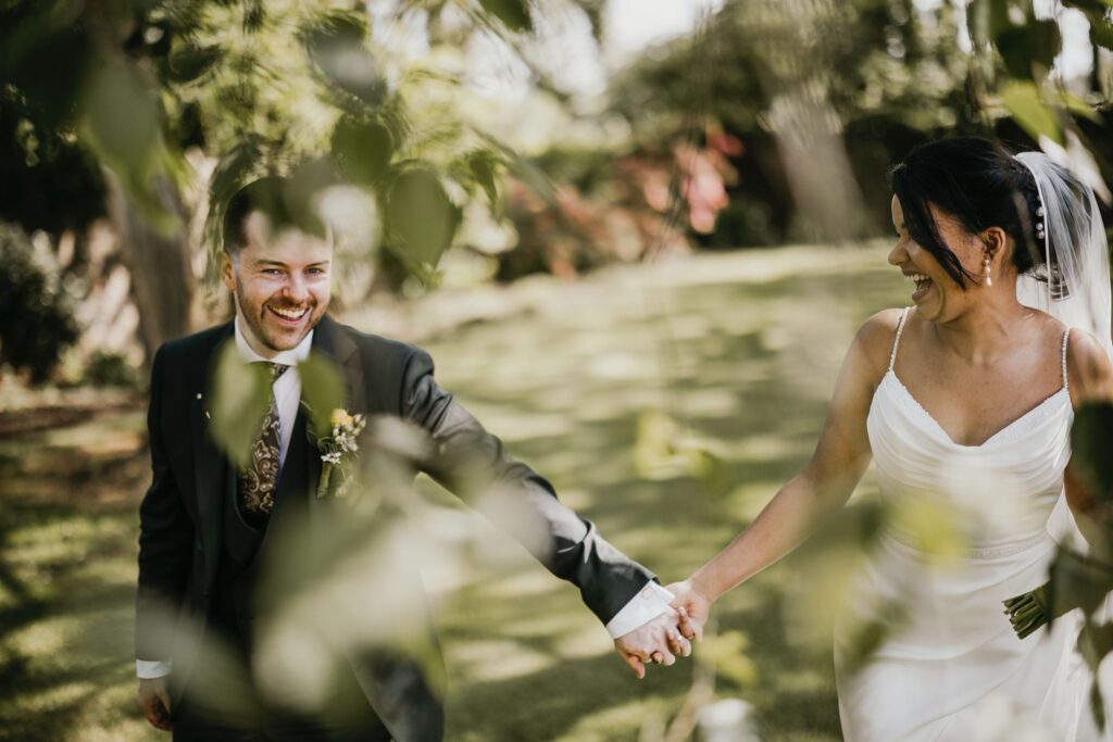 Bride and groom smiling at each other through green foliage.