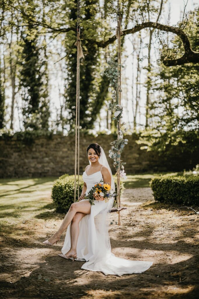 Bride in white dress swings on a floral-adorned swing beneath leafy trees