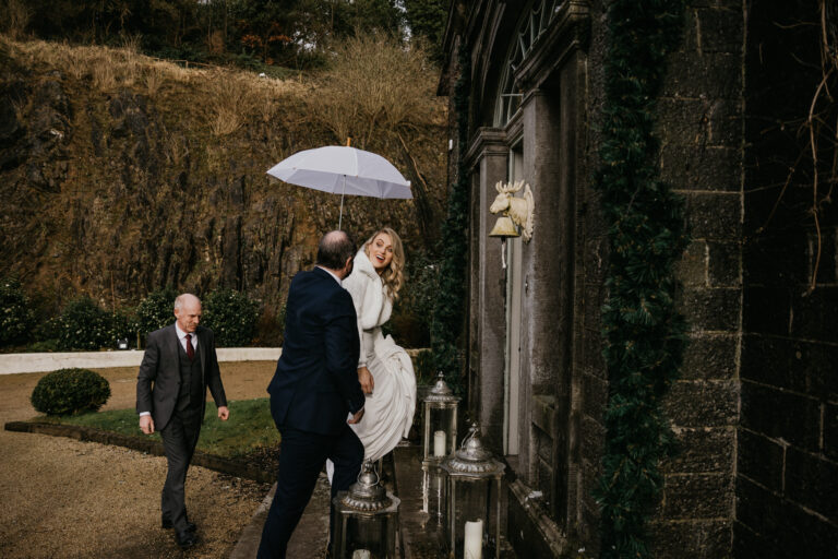 Bride standing under an umbrella at the door of a hotel, with the umbrella held by a porter – captured by Darren Byrne, Wedding Photographer in Ireland.