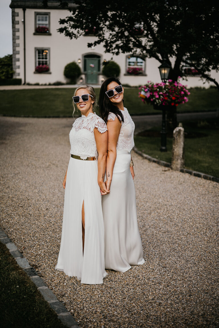 Two brides standing back to back outside wearing sunglasses and smiling