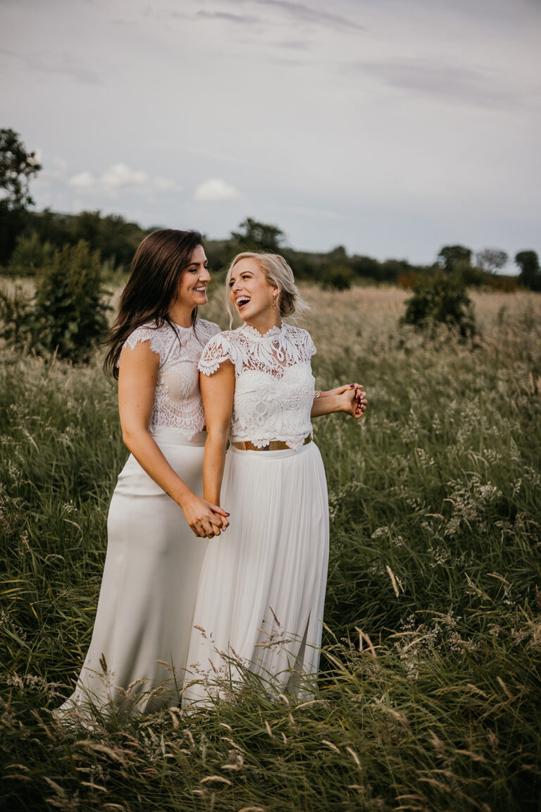 Two brides holding hands and glancing at each other in a meadow