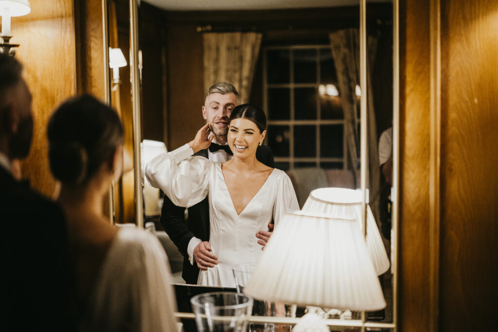 Bride holds groom’s face while smiling and looking in the mirror